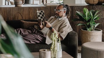 A person sitting on a couch in a room with a wooden cabinet, a table with a checkered object, and several potted plants.