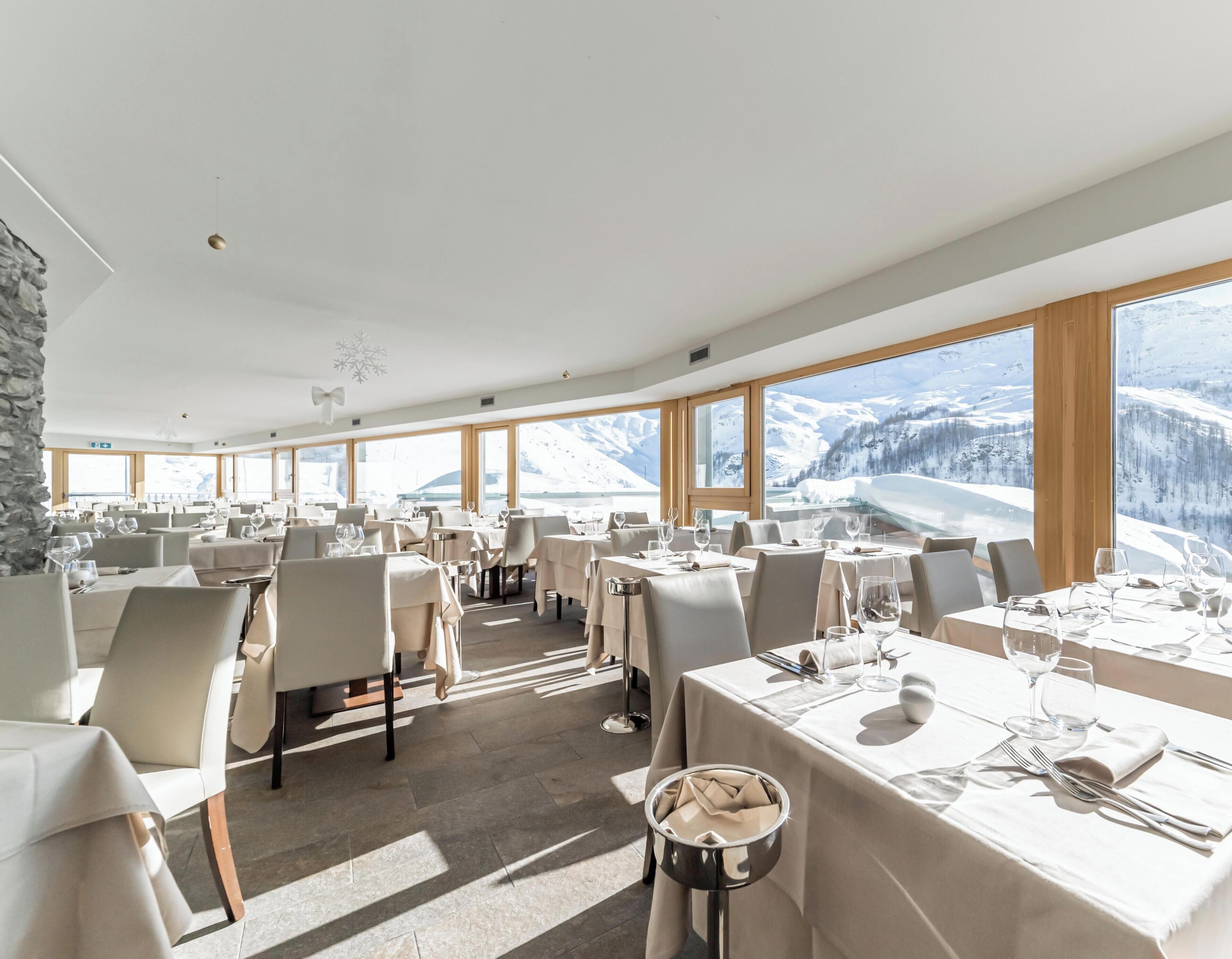 A restaurant with multiple tables set for dining, large windows offering a mountain view, and a stone wall on one side.