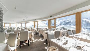 A restaurant with multiple tables set for dining, large windows offering a mountain view, and a stone wall on one side.