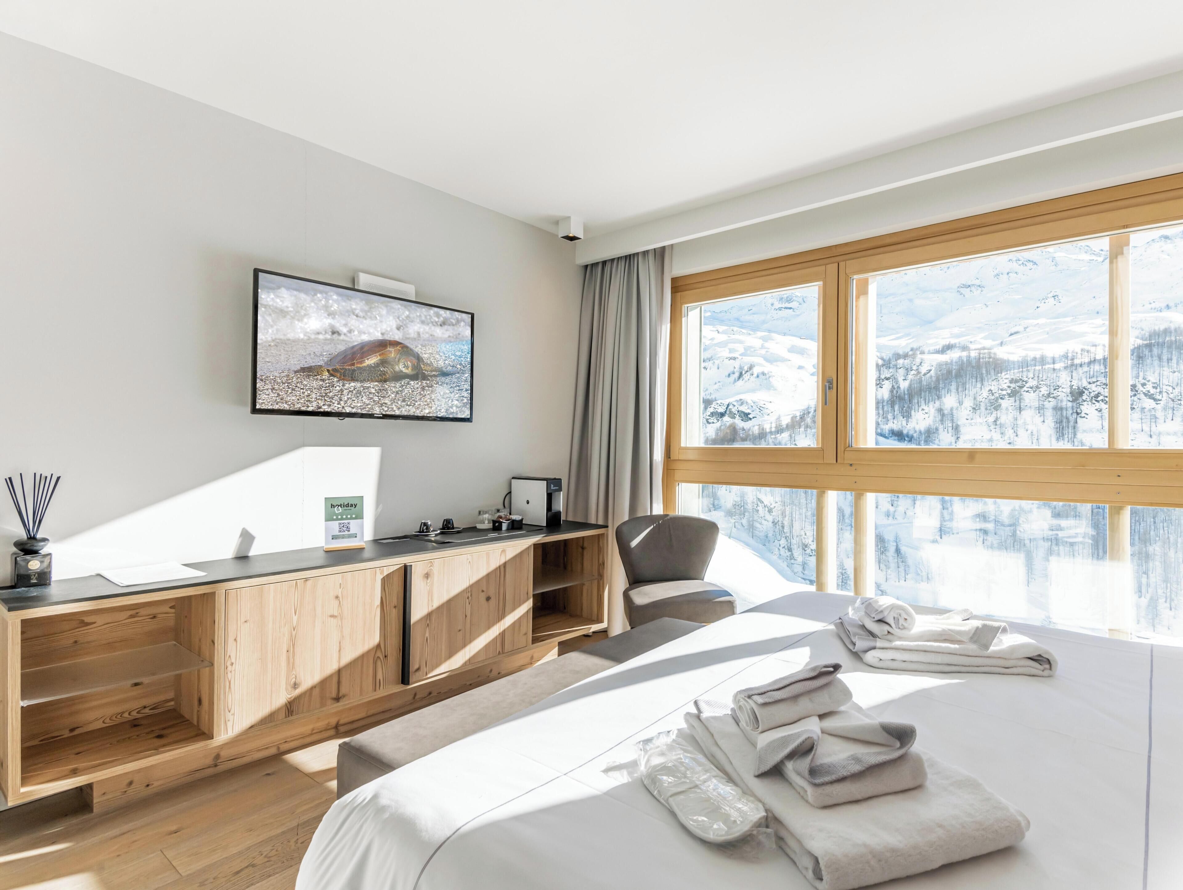 Une chambre d’hôtel moderne avec un grand lit, un bureau en bois, une chaise, un téléviseur à écran plat et une fenêtre donnant sur une vue enneigée sur les montagnes.