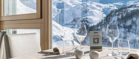 Mesa lista para comer con vistas a la montaña, un letrero con código QR y una ventana con panorama de montañas nevadas.