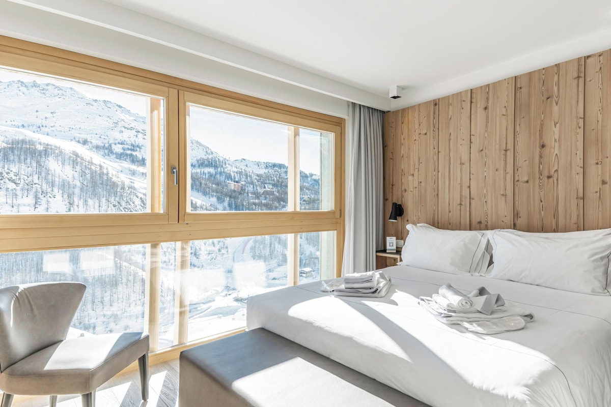 A modern hotel room with a large bed, a chair, and a view of a snowy mountain landscape.