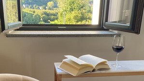 A room with a view of a green landscape through a window, a wooden table with an open book and a glass of wine, and a beige armchair. - Cascina 1909 (Priocca)