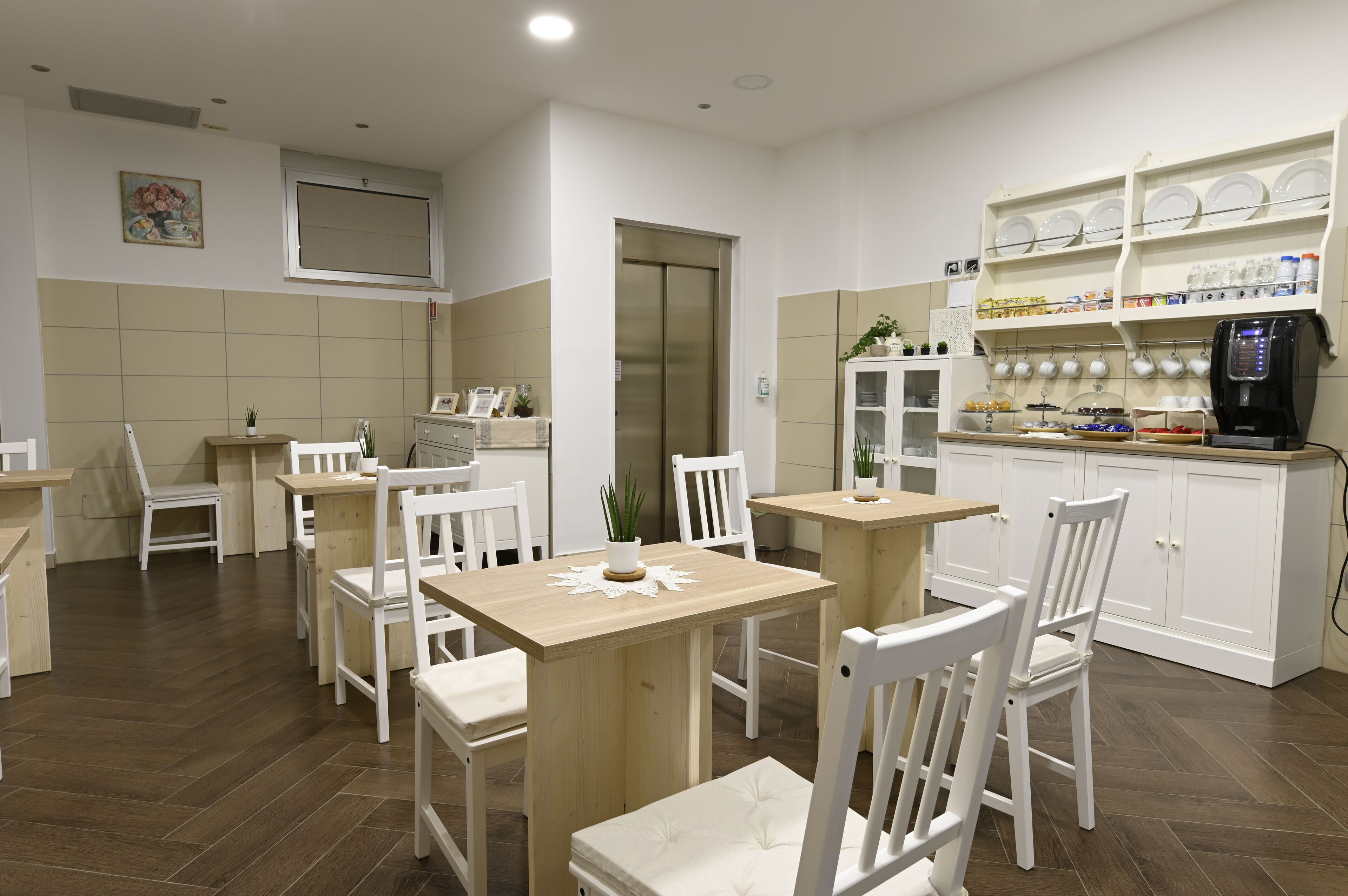 A dining area with wooden tables and white chairs, a kitchen with white cabinets and shelves, and a coffee machine.