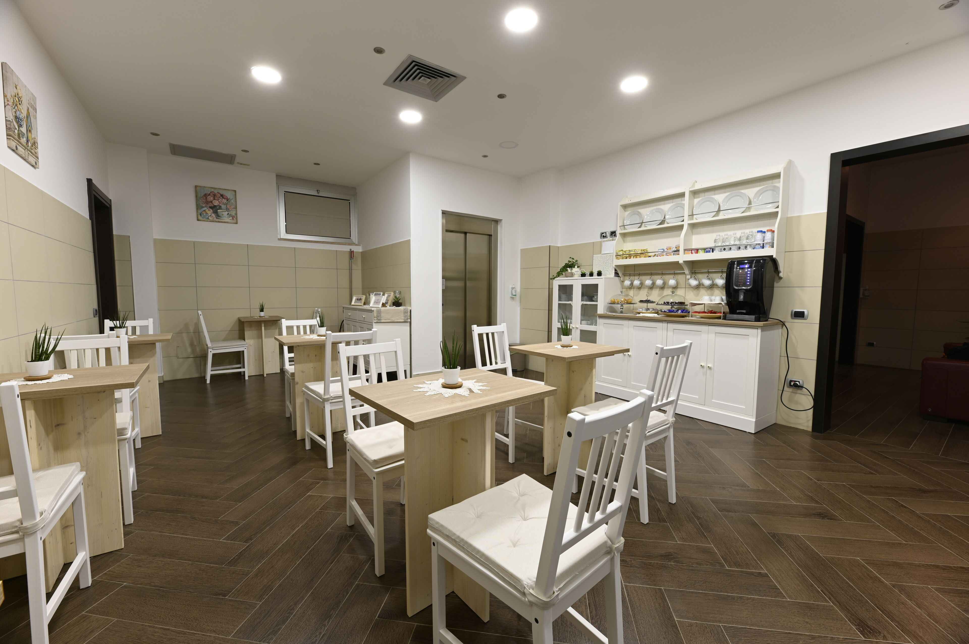 A dining area with white chairs and tables, a kitchen area with shelves, and a coffee machine.