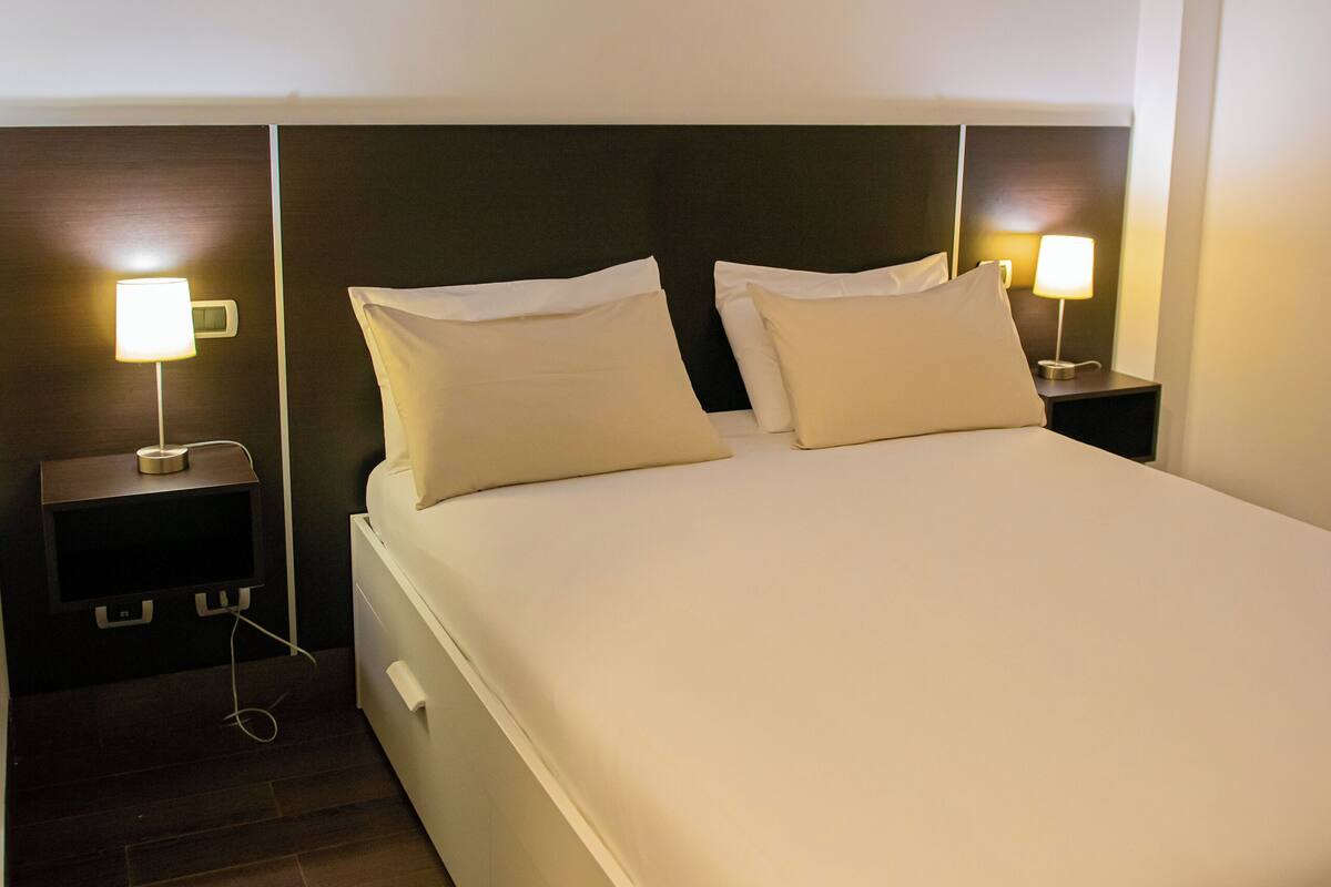 A neatly made bed with white linens and pillows, two bedside tables with lamps, and a wooden floor.