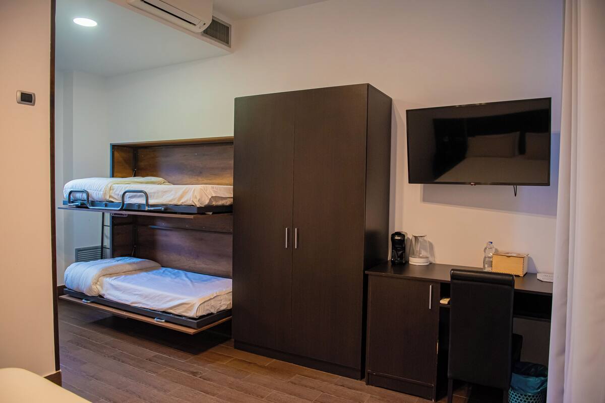 A compact hotel room with a bunk bed, a wardrobe, a desk with a chair, and a flat-screen TV mounted on the wall.