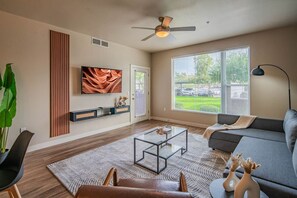 A modern living room with a flat-screen TV, a ceiling fan, a large window with a view of a green lawn, and a gray sofa. - Prime Location with Balcony, Pool, Gym, WD, Parkin (Tempe)