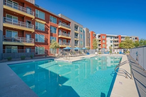 A modern apartment building with a swimming pool, outdoor seating, and a clear blue sky.
