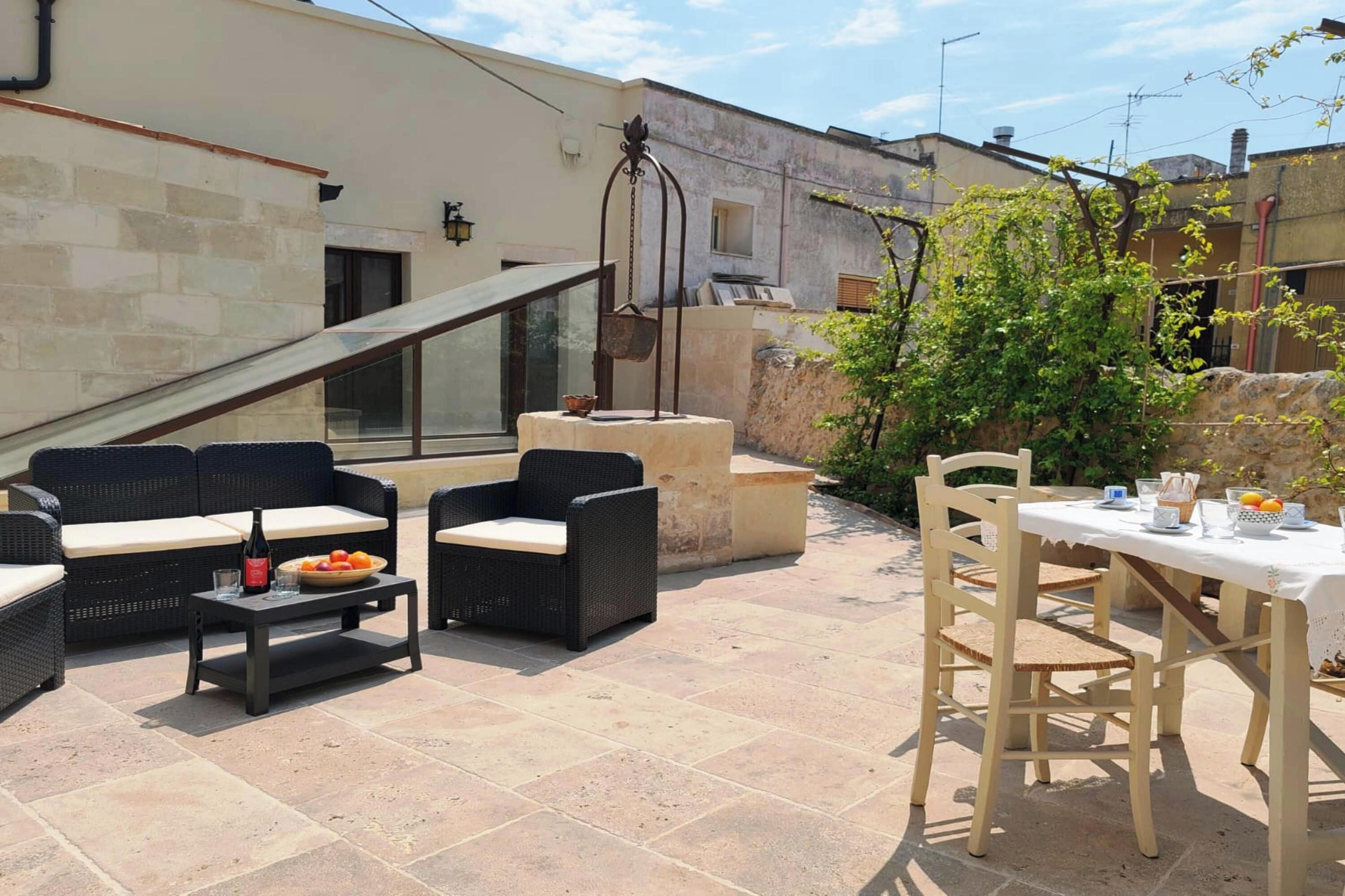 Un patio exterior con mobiliario de mimbre, una mesa dispuesta para comer y un pozo de piedra.
