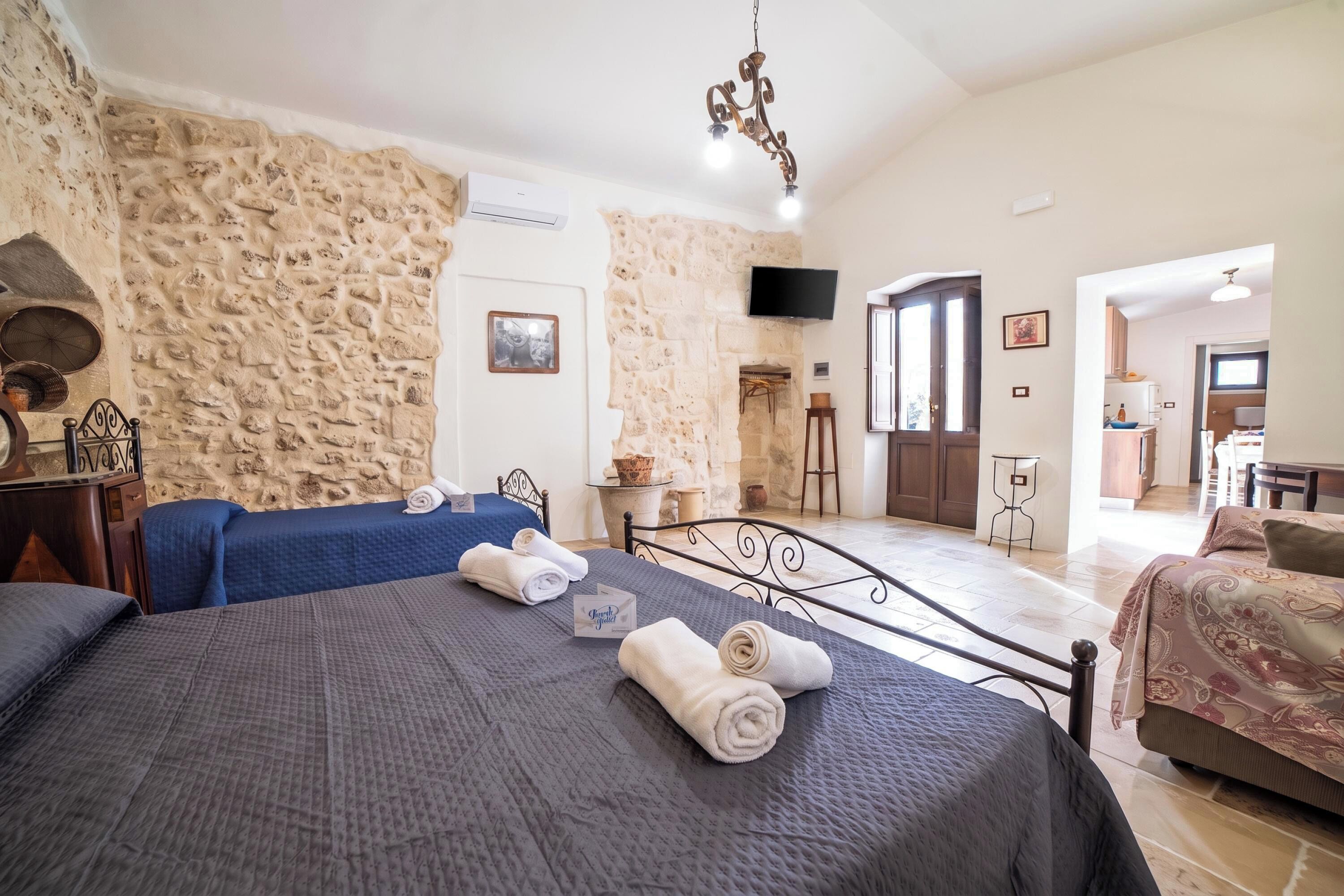 A hotel room with a stone wall, a bed with a blue bedspread, a television, and a dining area visible through an open door.