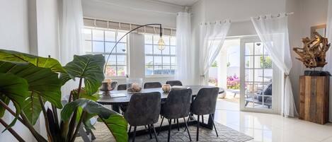 Uma sala de jantar com uma mesa posta para quatro pessoas, rodeada por cadeiras, e grandes janelas com cortinas.