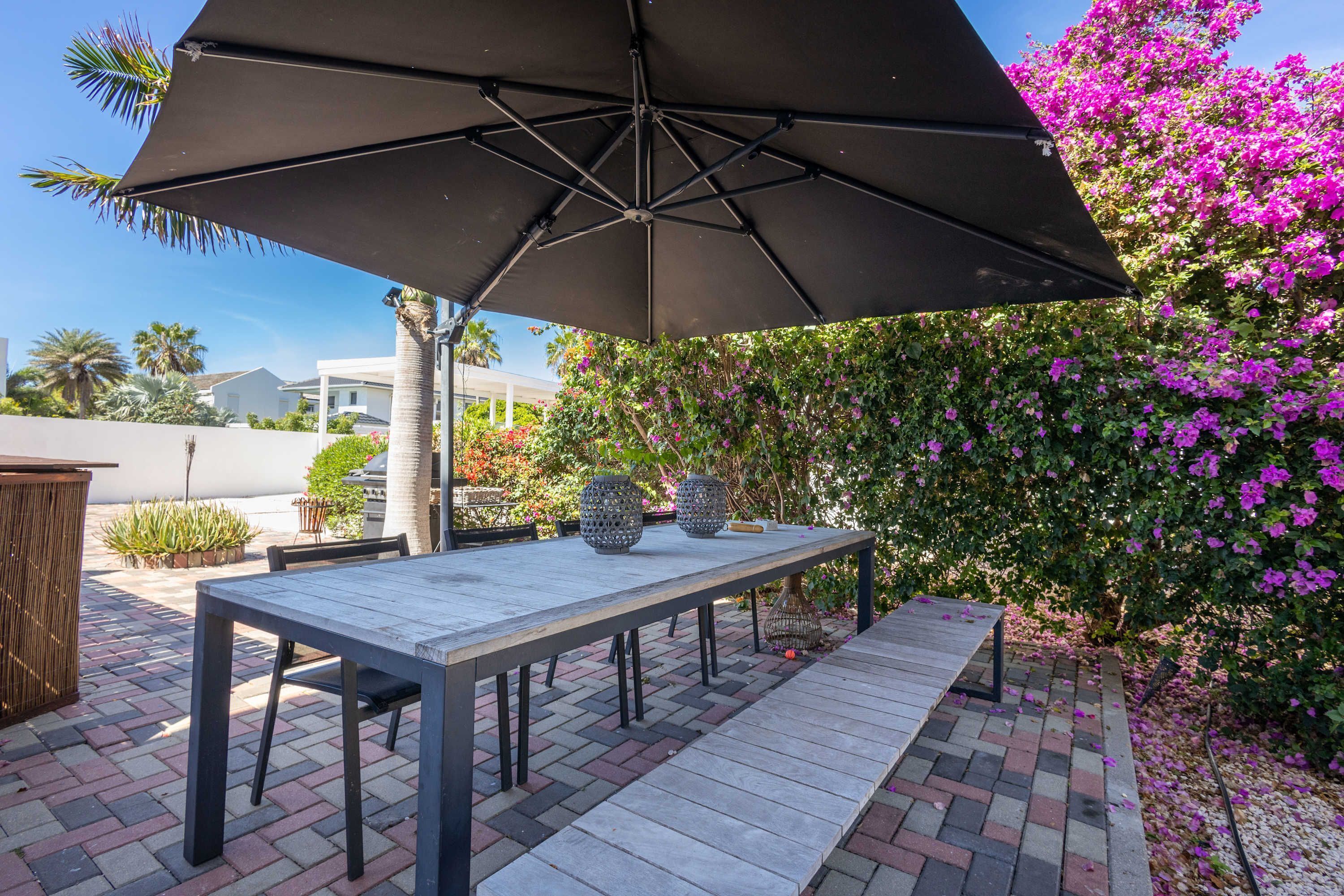 An outdoor patio with a table and chairs under a large umbrella, surrounded by potted plants and flowering bushes.