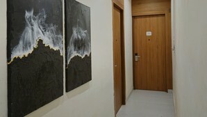 A hallway with two black and white abstract paintings on the wall, a wooden door at the end, and a ceiling light. - The Royal Orchid (Aligarh)