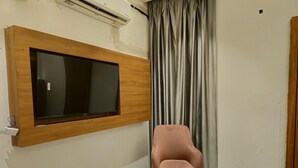 A room with a mounted TV, a pink chair, a curtain, and a wooden wall panel. - The Royal Orchid (Aligarh)