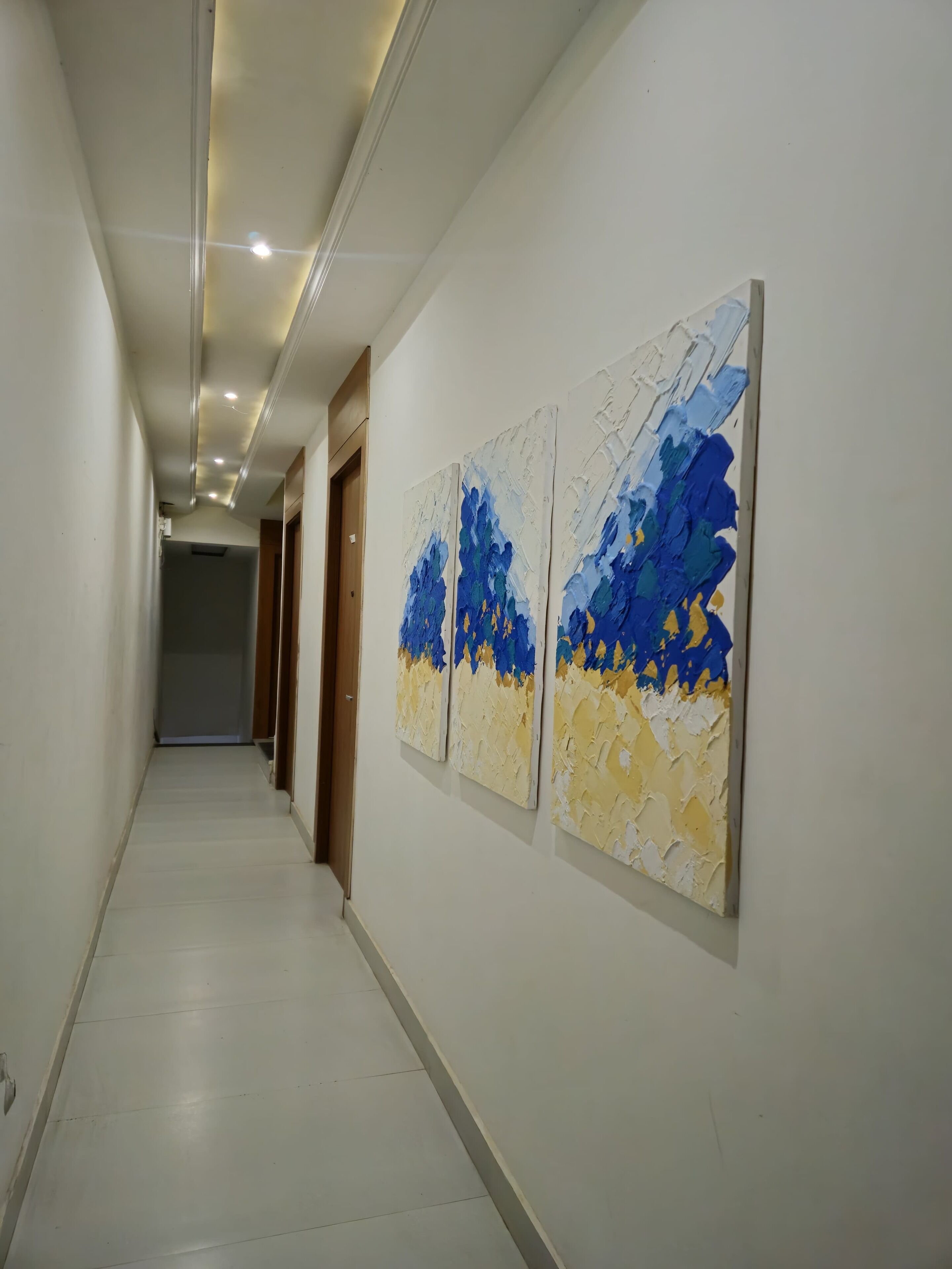 Un couloir aux murs blancs, doté d’un sol carrelé et orné de trois tableaux abstraits sur le mur de droite.