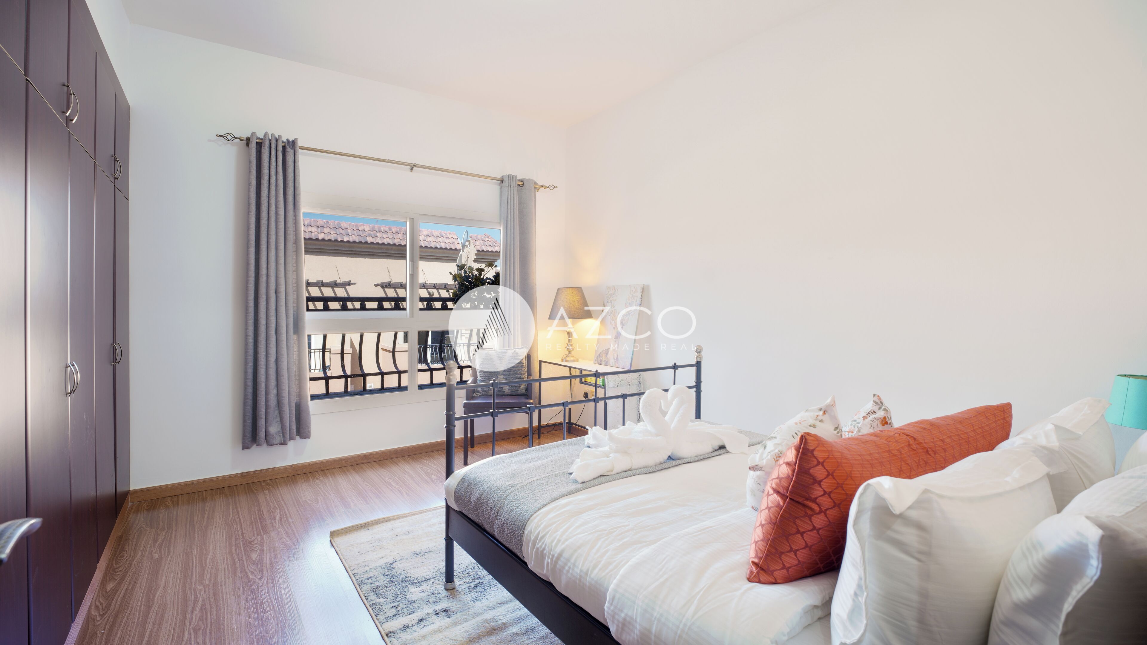 A bedroom with a bed, a window with curtains, a balcony, and a view of buildings.