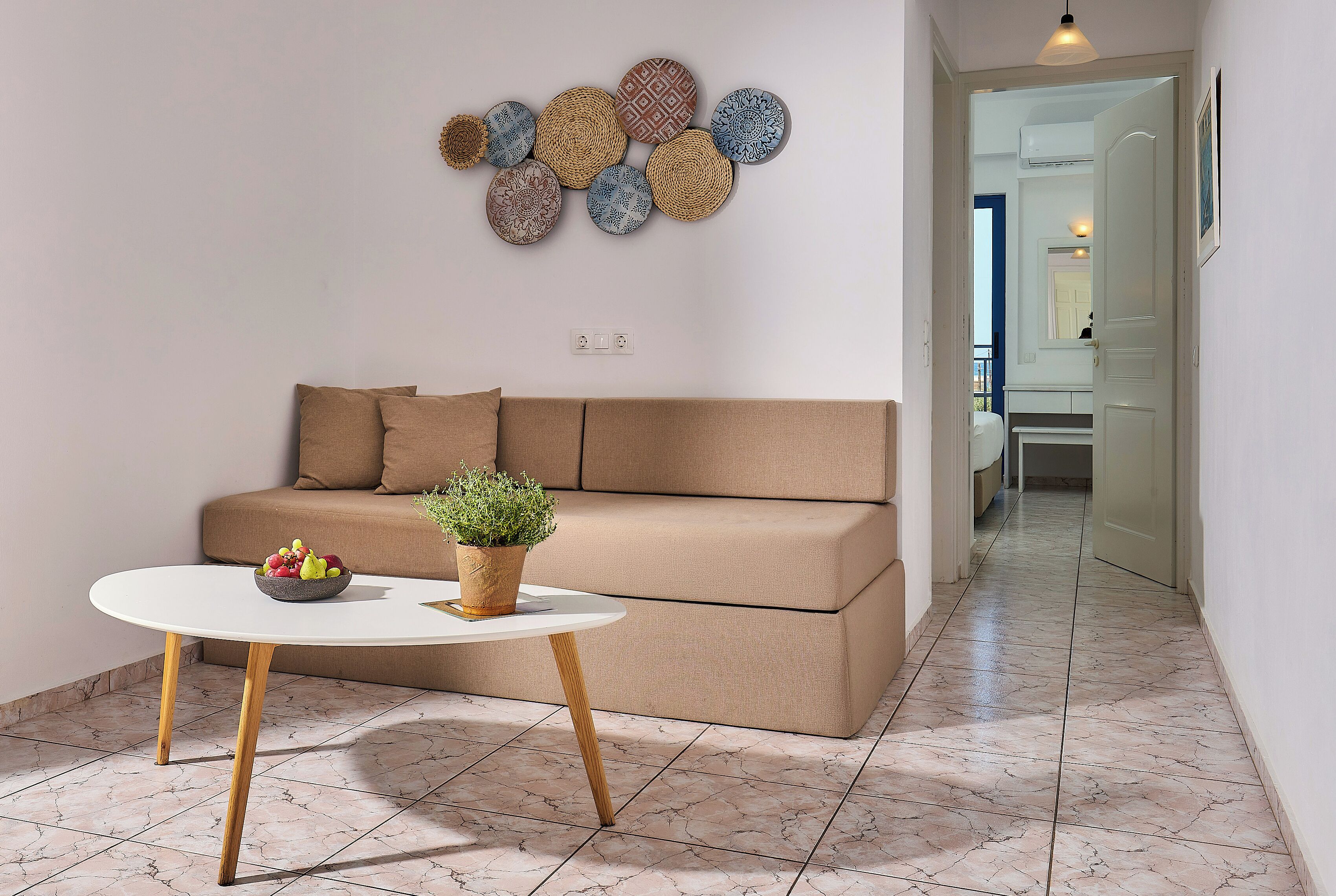 A modern living room with a beige sofa, a round white table with a bowl of fruit and a potted plant, and a wall decorated with circular woven art pieces.