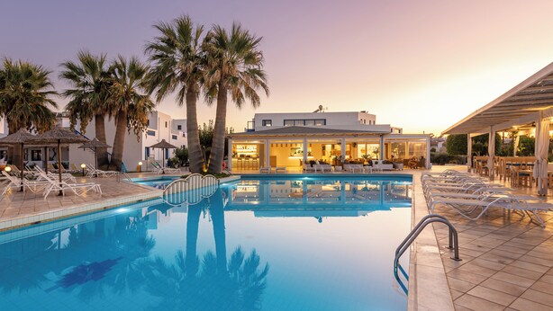 A swimming pool with lounge chairs and umbrellas, a bar area, and a building with large windows. - Ilios Suites (Hersonissos)