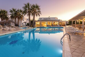 A swimming pool with lounge chairs and umbrellas, a bar area, and a building with large windows.
