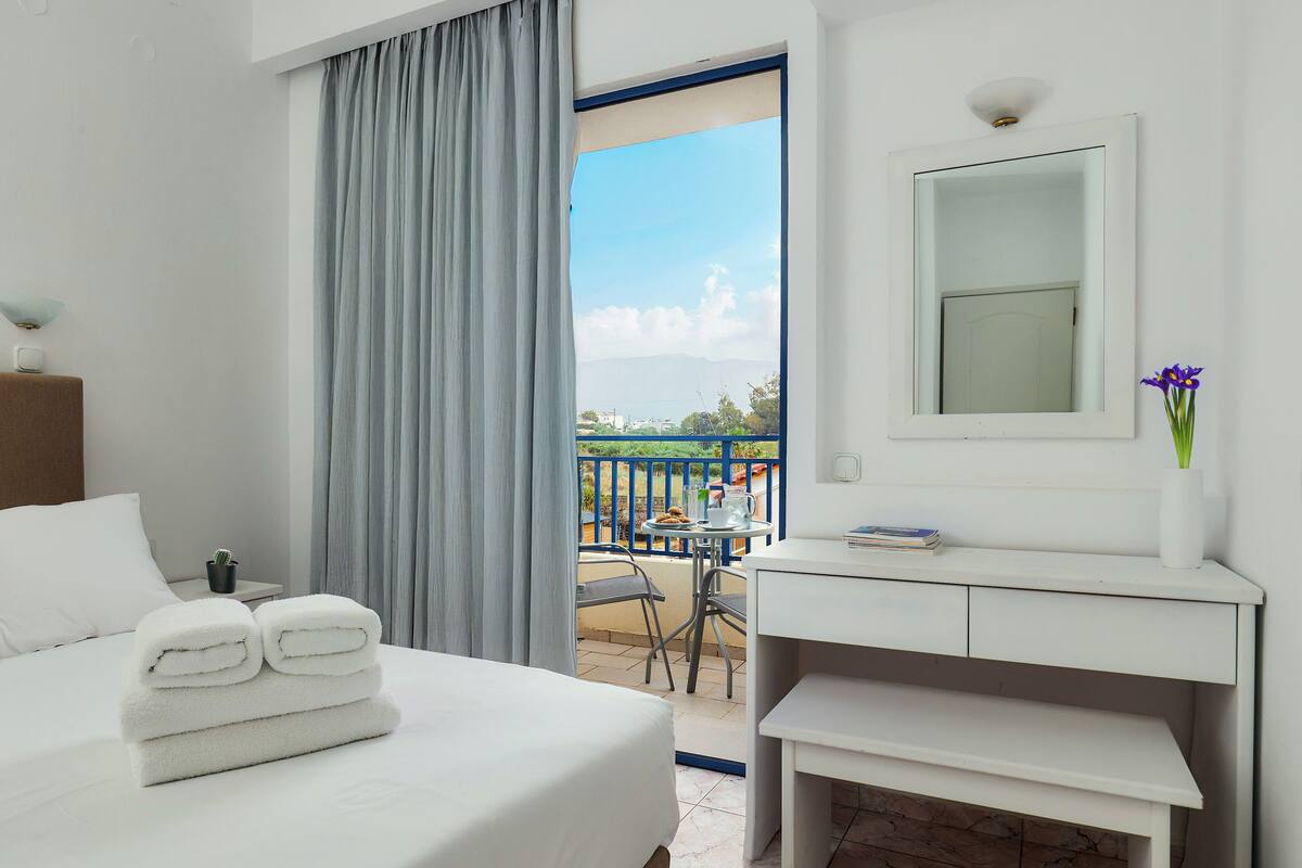 A hotel room with a bed, a desk, a chair, a small table with a plant, and a view of the outdoors.