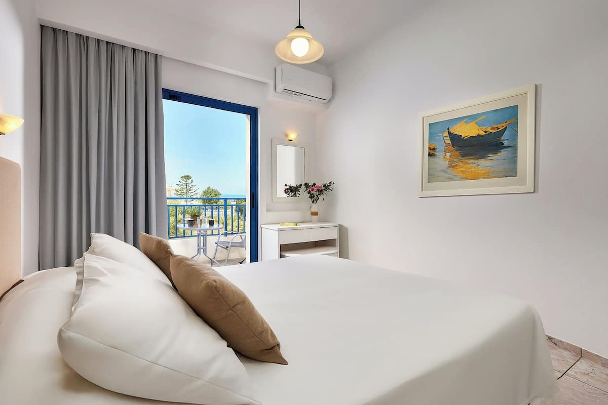 A hotel room with a bed, a desk, a chair, a balcony with a view, and a painting on the wall.