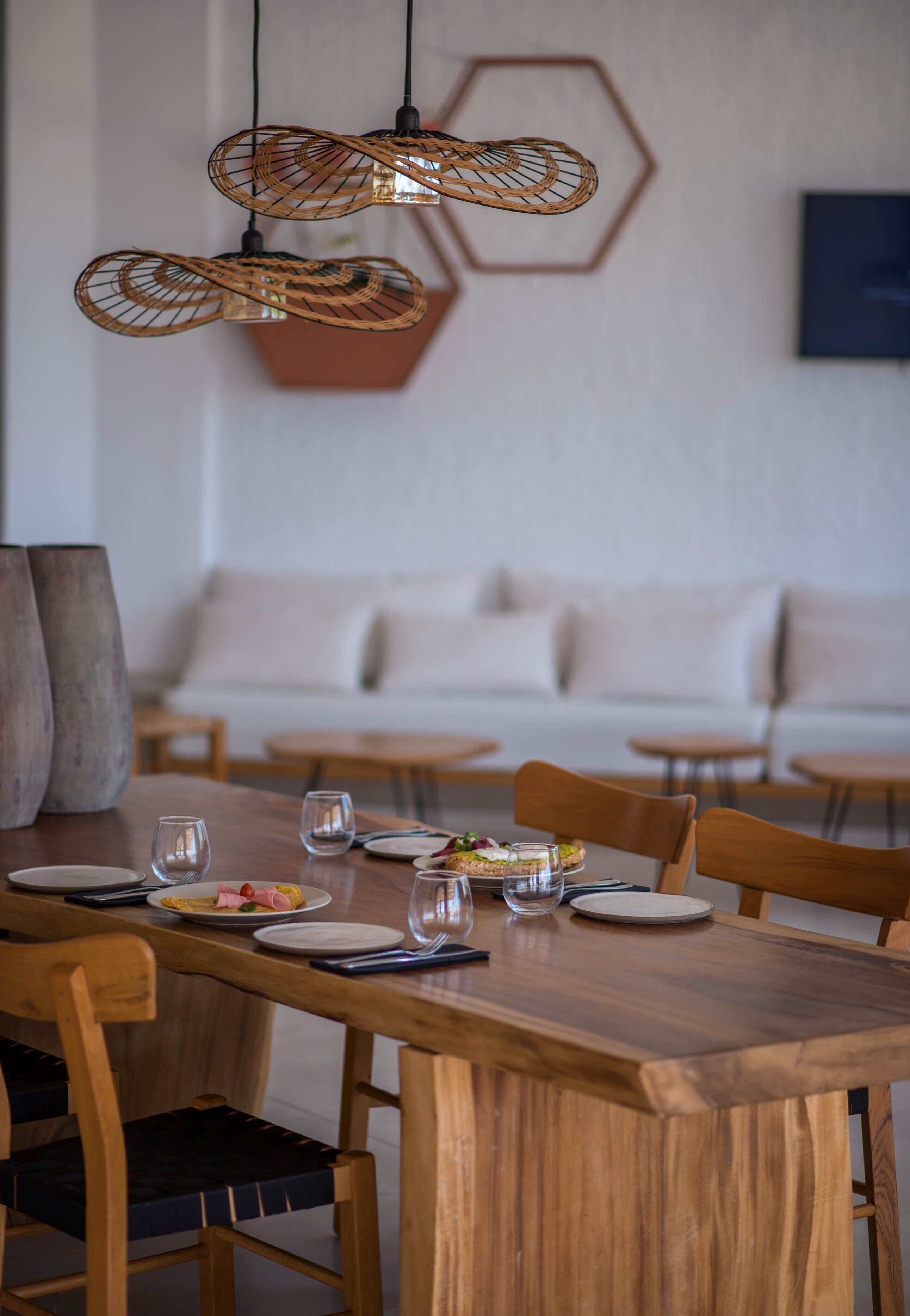 Een eetgedeelte met een houten tafel gedekt voor twee personen, een geweven hanglamp en een bank op de achtergrond.
