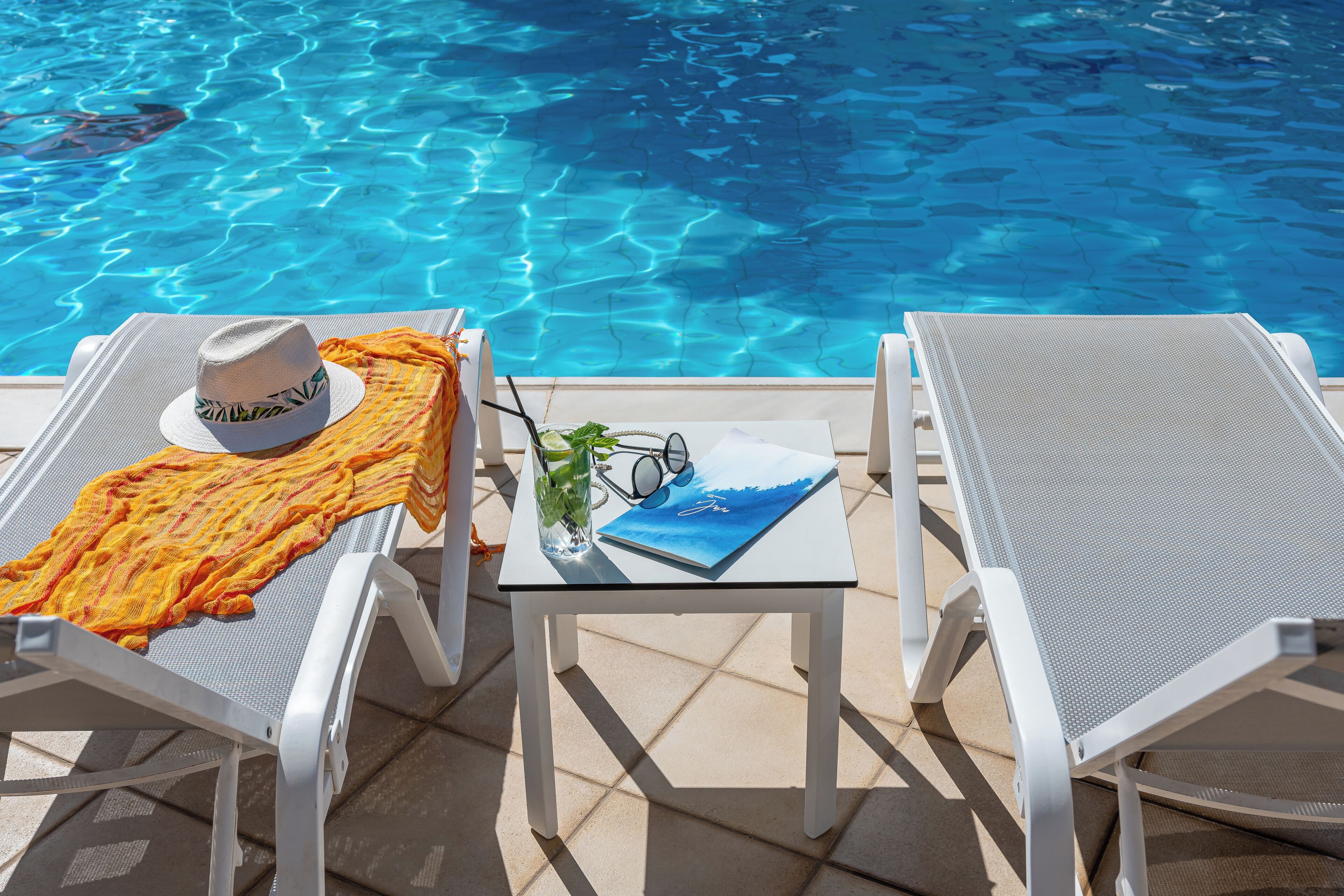 Deux chaises longues sont placées au bord d’une piscine. Sur l’une, on trouve un chapeau et un livre, et sur l ’autre, un verre d’eau et des lunettes de soleil.