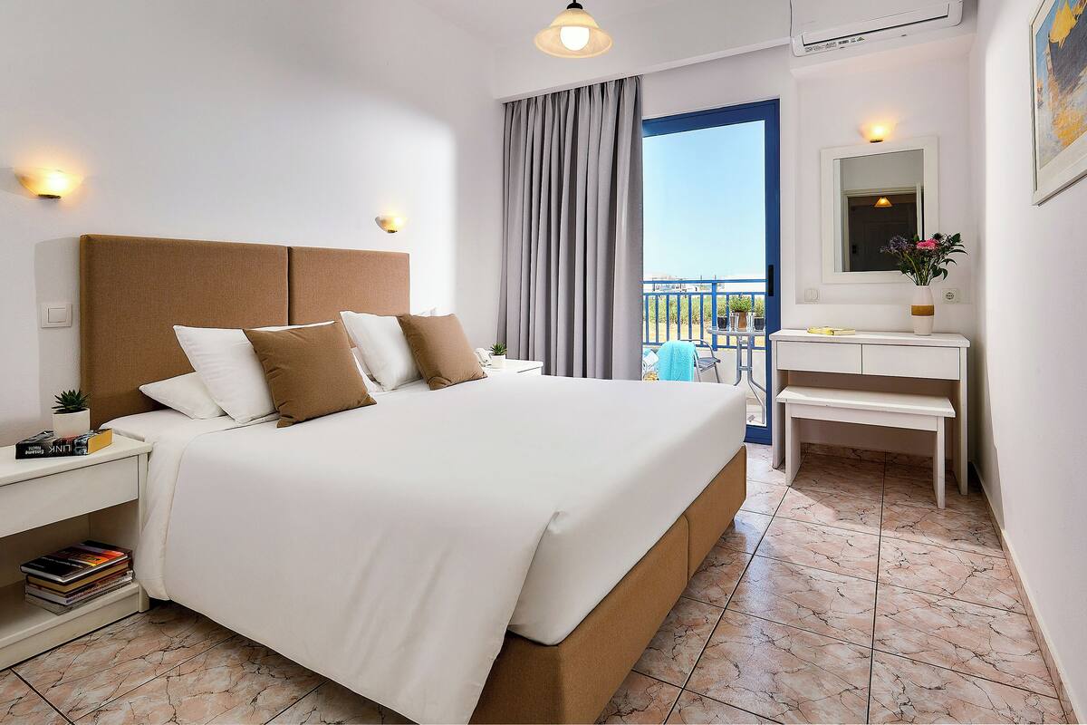 A hotel room with a large bed, bedside tables, a small desk, a mirror, and a balcony view.