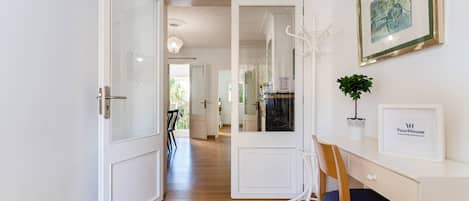 A hallway with a wooden floor, white walls, and a door leading to another room. There is a framed picture on the wall and a small potted plant on a table.