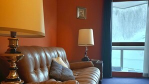 Ett rum med en brun lädersoffa, ett träbord med en lampa och ett fönster med utsikt över omgivningen.