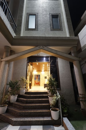 A building entrance with a staircase leading to a brightly lit interior, potted plants, and a metal railing.