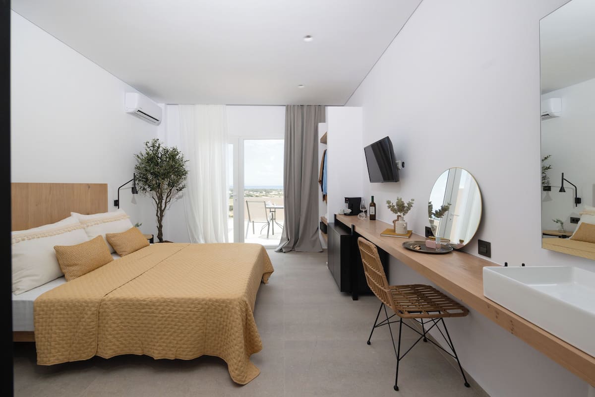 Quarto de hotel moderno com uma cama grande, uma escrivaninha com cadeira, pia, espelho e televisão.