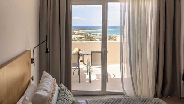 Un dormitorio con una cama grande, vistas a la playa y un balcón con mesa y sillas.