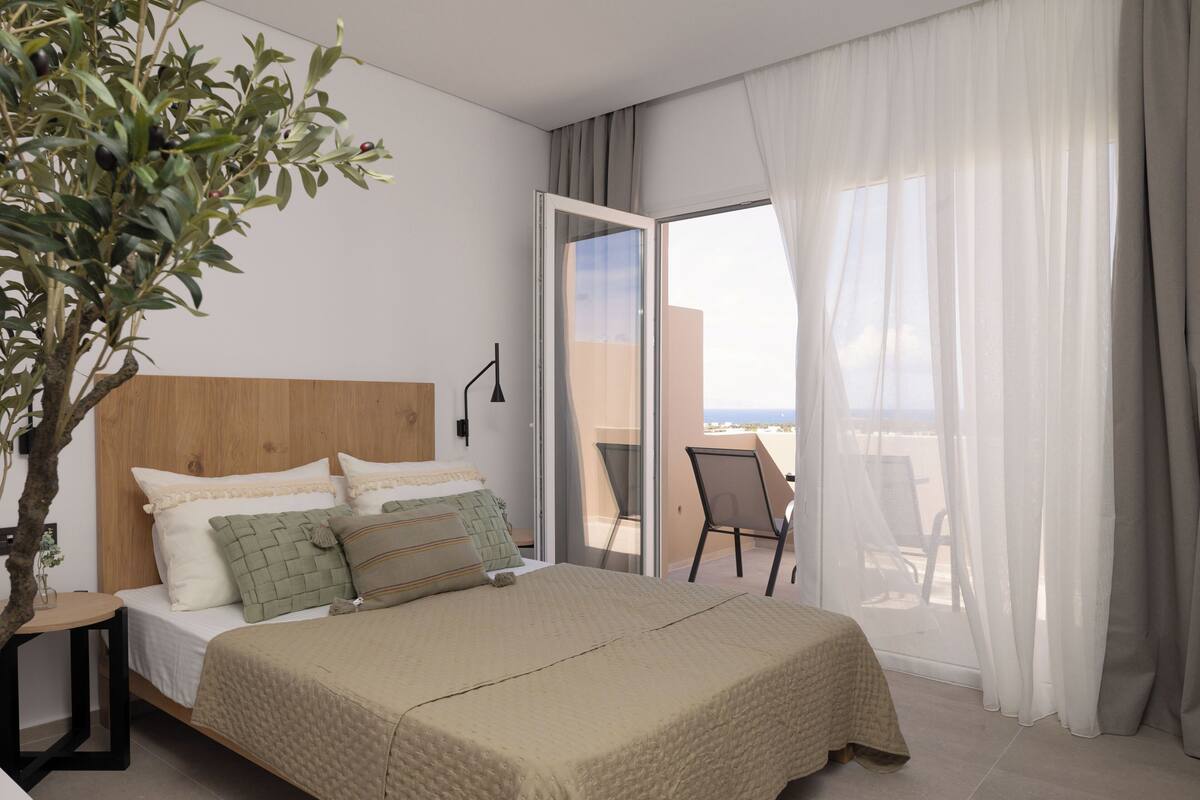 Quarto com cama, planta em vaso, varanda com cadeira e vista do céu.