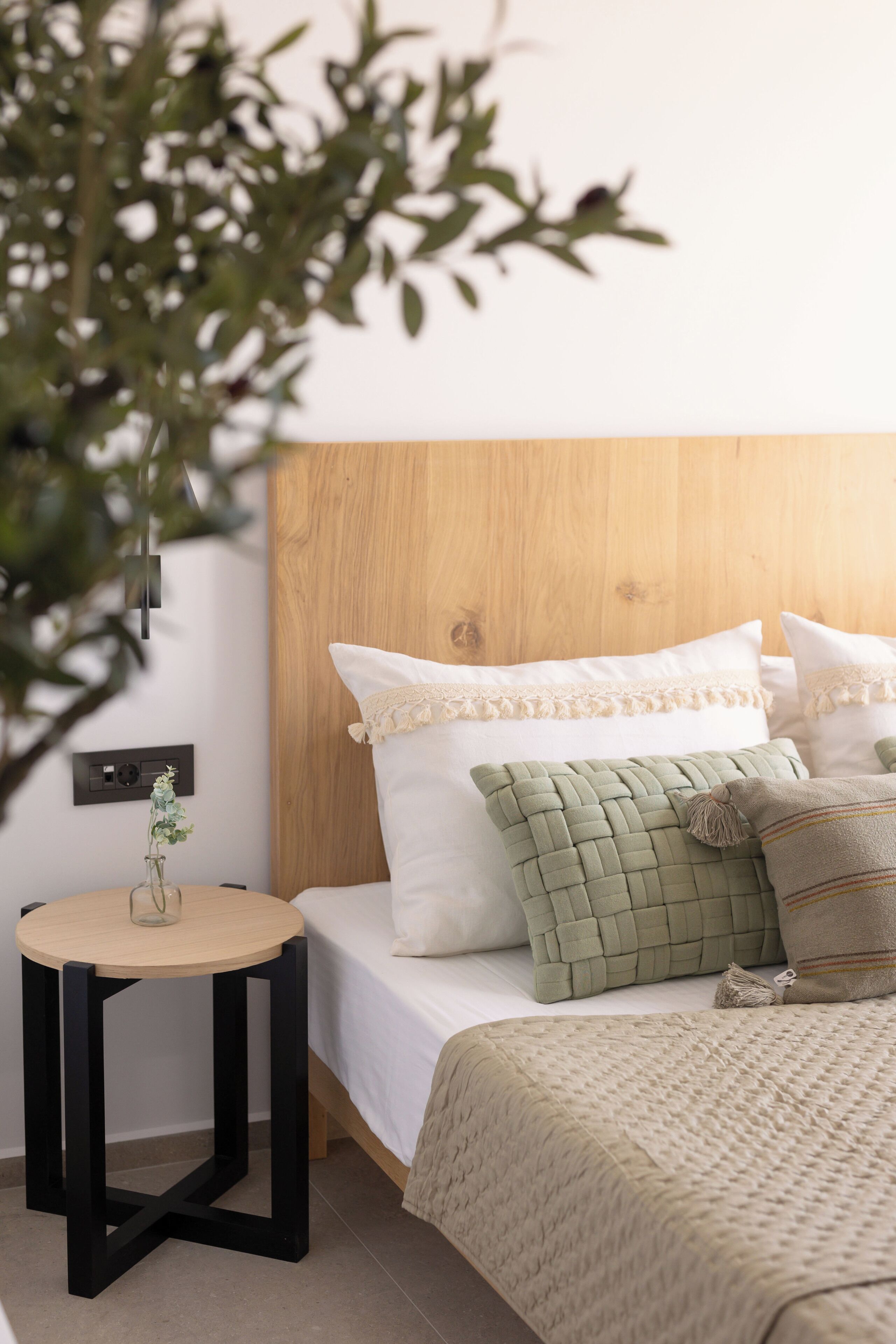 A neatly made bed with white and green pillows, a wooden headboard, and a small round table with a vase of flowers.