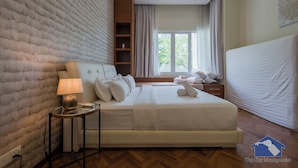 A modern hotel room with a large bed, a bedside table with a lamp, a window with curtains, and a view of greenery outside. - The Suites Straits Quay (George Town)