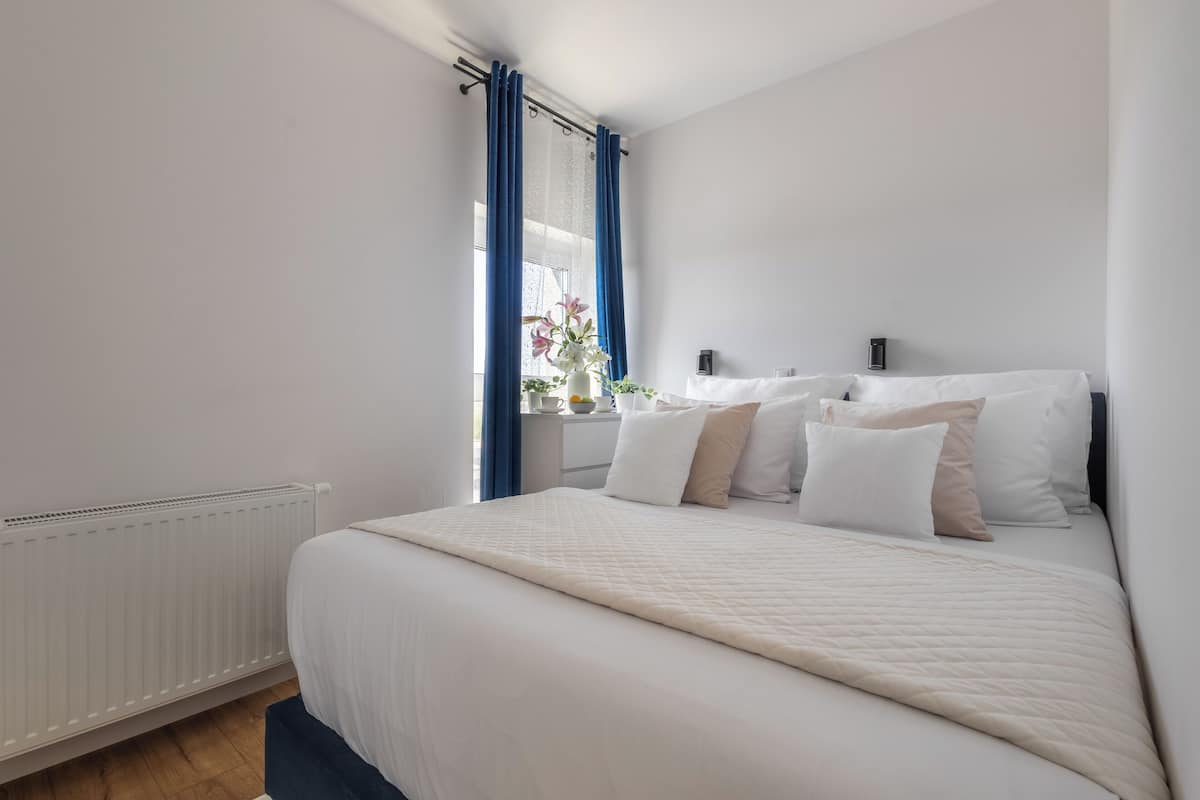 Un dormitorio con una cama grande, ropa de cama blanca, una ventana con cortinas azules y un radiador blanco.