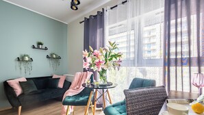 Una sala moderna con un sofá negro, sillones color aguamarina, una mesita redonda con un jarrón de flores y una ventana con cortinas de gasa.