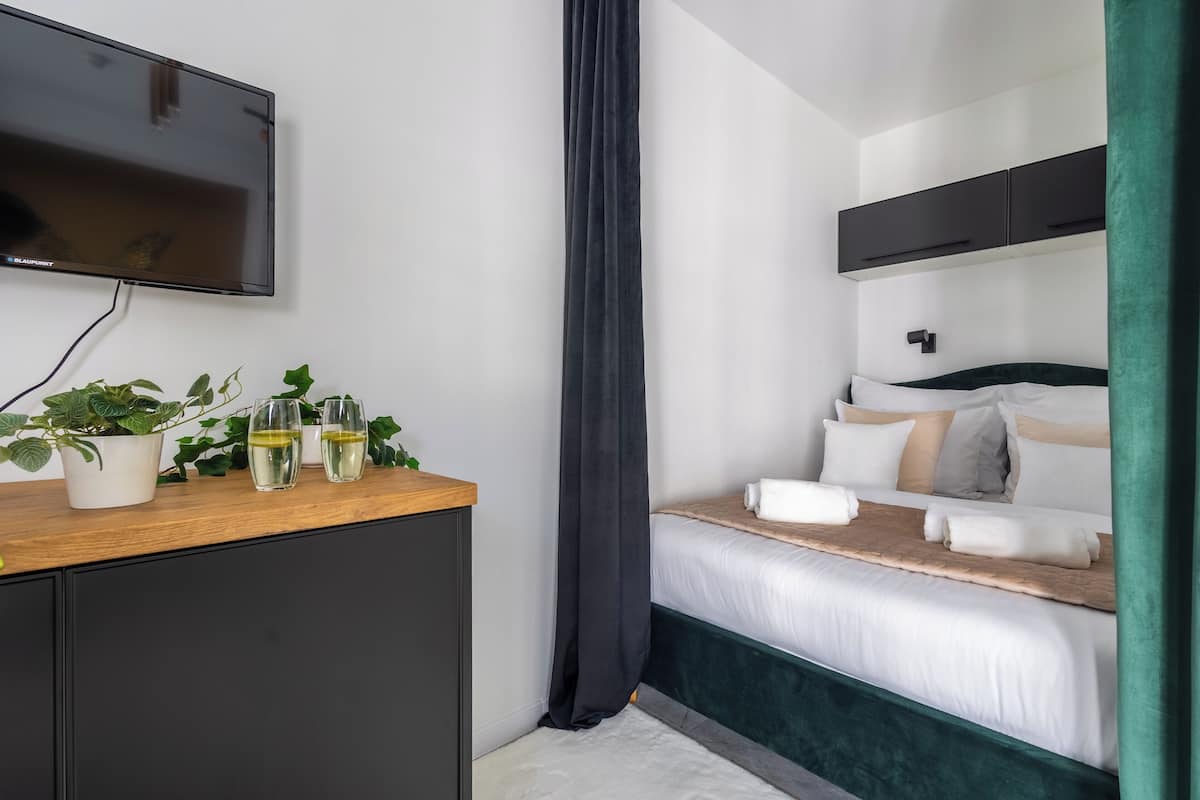 Una habitación de hotel moderna con una cama, un televisor montado en la pared, una mesita de madera con plantas y vasos, y una cortina verde.