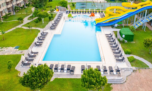 Una piscina rectangular grande rodeada de sillones de descanso y sombrillas, con un tobogán y un pequeño espacio verde cerca.