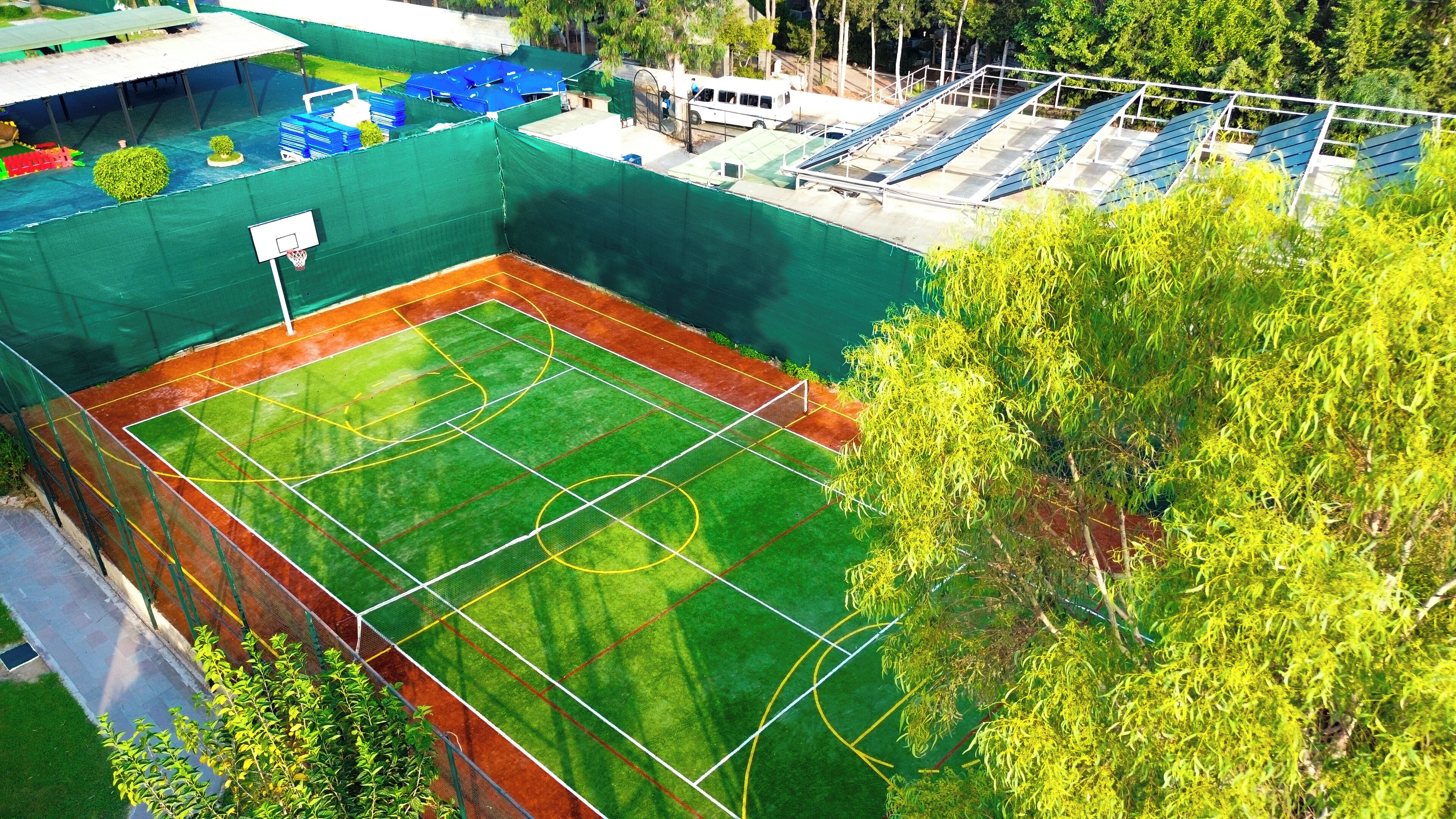 An outdoor tennis court surrounded by green fencing, with solar panels and a white vehicle in the background.