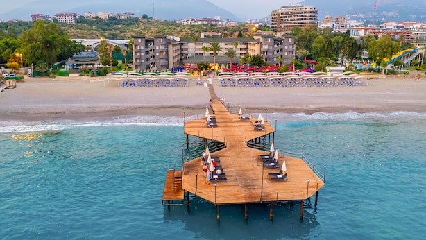 Un muelle de madera con sillones de descanso y sombrillas que se adentra en un mar azul cristalino, bordeado por una playa y edificios al fondo.
