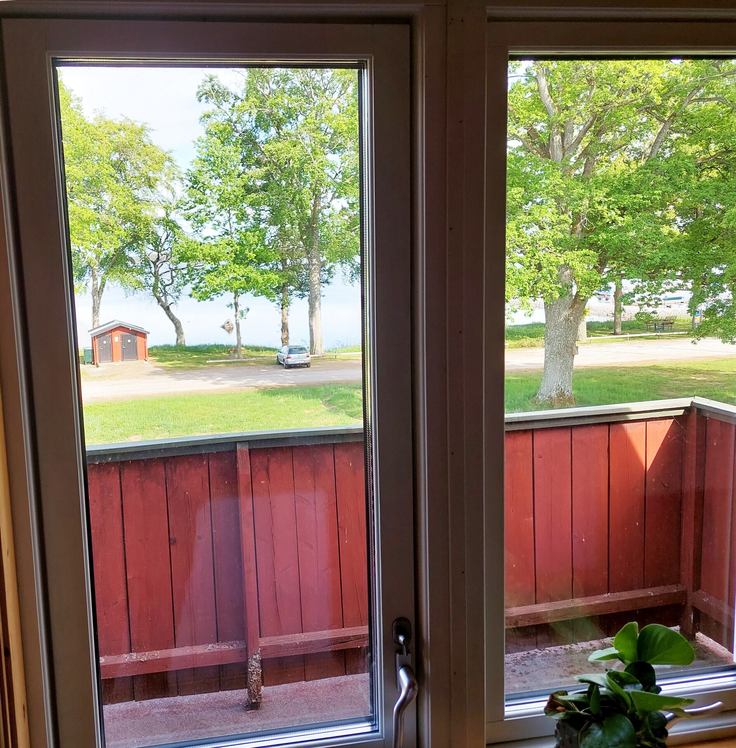 Une vue à travers une porte en bois dotée d’une poignée en métal, qui donne sur une terrasse en bois rouge et une cour verdoyante avec des arbres.