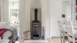 Ein geräumiges Wohnzimmer mit Holzboden, Kamin, Essbereich mit weißen Stühlen und einem großen Fenster, das viel natürliches Licht hereinlässt.