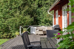 Relax on the deck surrounded by lush greenery, with a barbecue grill and wicker seating for outdoor dining or relaxation.