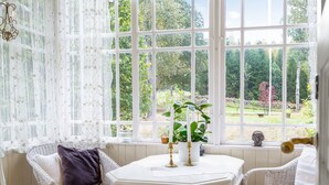 Ein gemütliches Zimmer mit Korbstühlen, einem weißen Tisch und Blick auf einen Garten durch große Fenster.