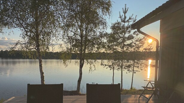 Puinen terassi, jossa on kaksi tuolia ja pöytä, ja josta avautuu näkymä tyyneen järveen, jonka rannalla kasvaa puita ja jossa auringonlasku värjää taivaan.