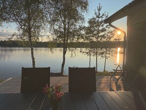 Un pontile in legno con due sedie e un tavolo, affacciato su un lago tranquillo, alberi e un tramonto.