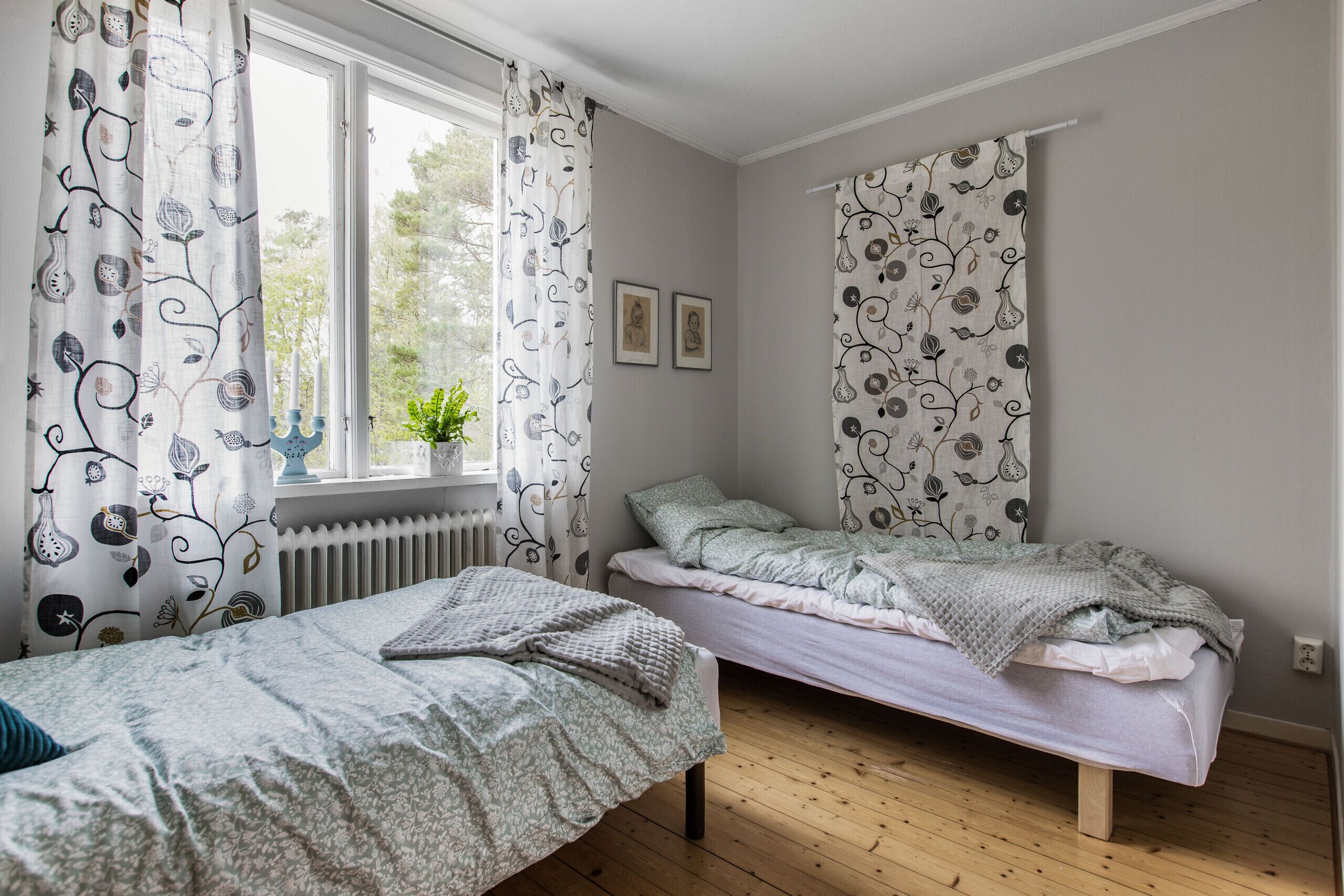 A bedroom with two single beds, a window with curtains, a radiator, and a potted plant.